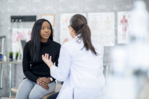 A woman of African descent and her doctor are indoors in a medical clinic. They are sitting and talking about the woman's health problems.
