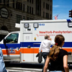 A NewYork-Presbyterian emergency medical services ambulance is stopped at a city intersection while pedestrians cross the street in a busy urban setting.